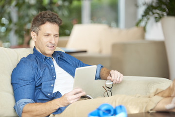 Wall Mural - Shot of a smiling confident man using his tablet while relaxing on the sofa at home.