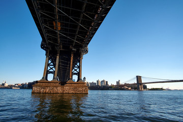 A closeup look up under the Manhattan Bridge near one of the supporting towers, with Brooklyn Bridge in the distance