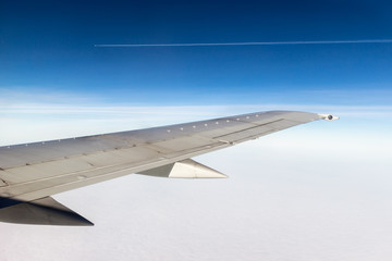 Wall Mural - Wing of a plane on blue sky background. Clouds underneath the wing. Parallel plane in a distance with trails