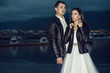 Young just married beautiful stylish couple in black leather jackets standing on the berth in the bay at dusk with sea and mountains on background. Outdoor shot. Copy-space