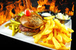 Plate with beef burger, bowl of white sauce, golden french fries, pickles and green vegetables. Studio shot with black wooden background and flames.