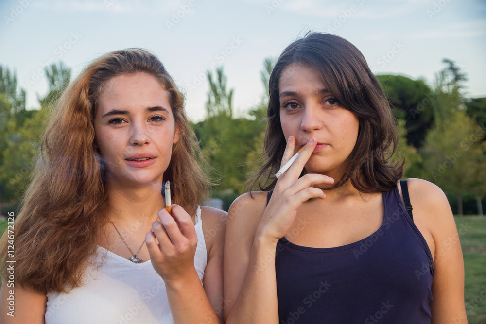 two women smoking cigarettes Two girls friends smoking cigarettes in the park on a cloudy day. They are  of celebration and are happy. Stock Photo