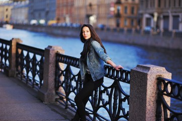 Pretty serious girl in a denim jacket standing on the St. Petersburg waterfront leaning against the iron railing and, looking into the camera.