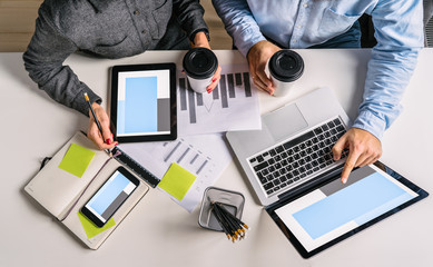 Wall Mural - View from above.Business people sitting at white desk. Businessman pointing at laptop screen,holding in his other hand cup of coffee.Teamwork.On table smartphone,digital tablet, graphics, notebook.