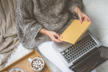 Sticker - Girl in beige knit sweater and white pants sitting at home in cozy atmosphere and is holding golden envelope.In lap of girl is laptop. Girl using gadget. In the background white knitted pillow.