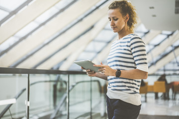 Wall Mural - Girl, dressed in striped jacket, standing in indoors and looking on screen digital tablet,located in her hands. Woman standing and holding in hands tablet computer.Girl uses digital gadget.Style life.