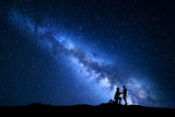 Fototapeta  - Milky Way. Night landscape with silhouettes of a man making marriage proposal to his girlfriend and starry sky. Silhouette of lovers. Couple, relationship. Blue Milky way with people. Universe