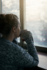 Sad and lonely woman looking out of the window and drinking coffee