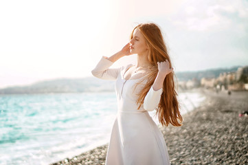 Outdoor summer portrait of young pretty woman with great hair looking to the ocean at europe beach, enjoy her freedom and fresh air, wearing stylish white dress.