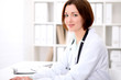 © rogerphoto - Young brunette female doctor sitting at the table and working at hospital office.  Health care, insurance and help concept. Physician ready to examine patient