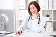 © rogerphoto - Young brunette female doctor sitting at a desk and working on the computer at the hospital office.  Health care, insurance and help concept. Physician ready to examine patient