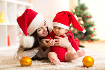 Lovely mother and baby in santa red dress smile on a background of Christmas trees in the interior of the house