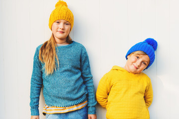 Wall Mural - Outdoor portrait of adorable fashion kids, wearing warm pullovers and hats, standing against white wooden background