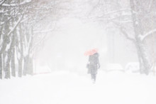 Silhouette Of A Woman In A Dark Coat With A Red Umbrella In A Snow Storm. Soft Focus, Blurred Outlines. Street Of The City In The Winter Blizzard And Snow With Beautiful Snow Flakes.