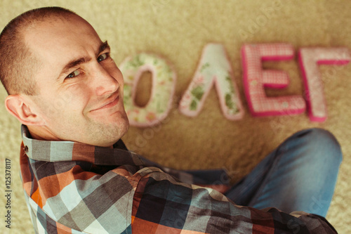 Fotografie, Obraz Funny adult man smiles while sitting over his name made of pillo