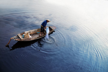 Asian fisherman on wooden boat casting a net for catching freshwater fish in nature river in the early morning before sunrise