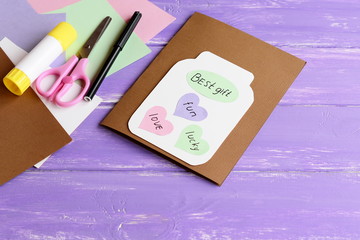 Wall Mural - Paper greeting card with jar, colored hearts and wishes. Scissors, colored paper sheets, white cardboard, glue stick, black marker on wooden background. Fun kids art concept