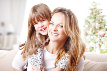 Mother and daughter at christmas. Close-up portrait of cute little girl hugging her happy mother while sitting at home in christmas time. 