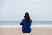 Lonely And Depressed Woman Sitting On The Sand Of A Deserted Beach And Watching The Sea On An Autumn Day.