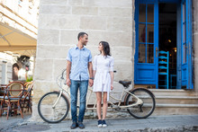 Guy And Girl Stand Holding Hands And Looking At Each Other On The Background Of Their Tandem Bike, Walls And Vintage Door On The Sidewalk Of A City Street. Lviv, Ukraine