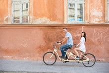 Happy Couple Riding On Retro Tandem Bicycle At The Street City Against The Background Of The Old Orange Wall With Windows. The Man Runs A Bicycle, A Girl In White Dress Raised Her Hand Up. Lviv