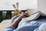 Fototapeta  - Young man reading a book lying on soft mattress in relaxing bed at terrace with green nature view. Fresh air in the morning of weekend or free day. Relax or education background idea. Selective focus.