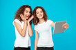 Two girls twins making selfie on tablet over blue background. 