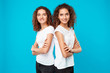 Girls twins posing with crossed arms, smiling over blue background.