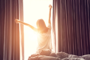 Woman standing near the window while stretching near bed after waking up with sunrise at morning, back view