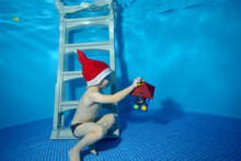 Child In Hat Santa Claus With A Toy In The Hand Swims Under The Water And Plays On The Bottom Of The Pool. The View From The Water Side. Horizontal Orientation