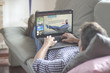 © GENETTICA - Young man lying comfortably on the sofa while using the laptop with video on demand website on the screen. View from behind. All screen graphics are made up.