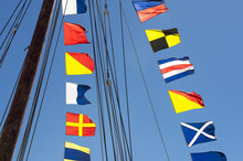 Colorful Nautical Sailing Flags Flying In The Wind From The Lines Of A Sailboat Mast Backlit In Bright Blue Sky By The Sun