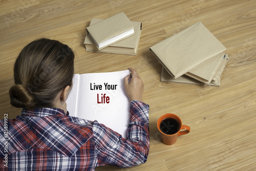 Fotografie, Obraz Woman reading book Live Your Life by stack of books
