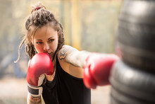 Woman Boxer With Tatto Arms At Workout. Young Woman Is Boxing Punching Bag From Car Tires. At The Head Of The Girl Dreadlocks. Toned Photo.