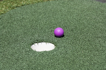 Purple golf ball beside hole on putt putt or miniature golf course