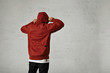 A young hipster adjusts the hood of his brownish red parka, back view, shot in studio with white walls