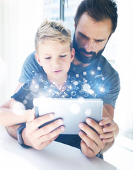 Bearded father with his young son using tablet PC in sunny room.Dad and little boy playing together on mobile computer, resting indoor.Childhood dreams icons concept.Vertical, blurred background.