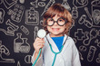 Happy little boy in doctor costume holding sthetoscope on dark background with pattern. The child has glasses