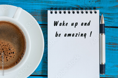 Poster Wake Up and Be Amazing message text written in notepad near morning cup of coffe
