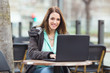 Busy young businesswoman is sitting in an outdoor café and using laptop. She is looking at camera and smiling.