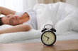 © rogerphoto - Alarm clock standing on bedside table has already rung early morning to wake up woman in bed sleeping in background. Early awakening, not getting enough sleep, oversleep, time line concept