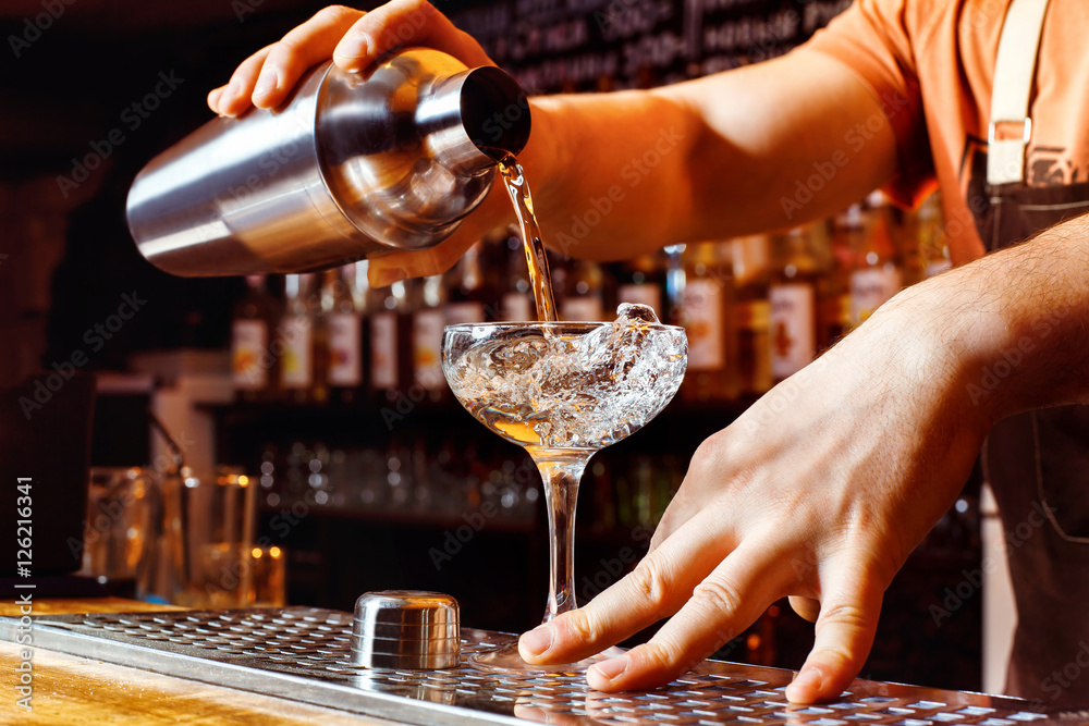 Male bartender is making cocktail holding shaker with pouring alcohol - obrazy, fototapety, plakaty 