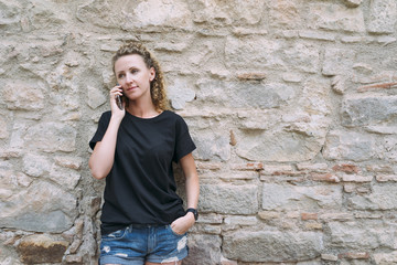 Wall Mural - Front view of a young woman dressed in a black T-shirt and denim shorts, standing and talking on the phone. In the background is bright stone wall. Girl uses the gadget.