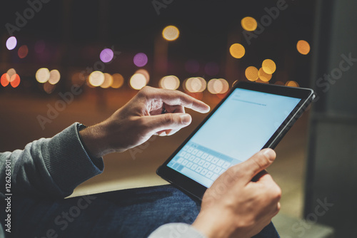 Fotografie, Obraz Side view of male hands using modern digital tablet outdoor, night lights and bo