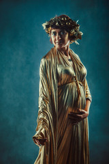 Pregnant woman in golden toga and wreath posing like a Greece fertility goddess