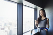 © Mint Images - A business woman by a window with a view over the city, using her smart phone.