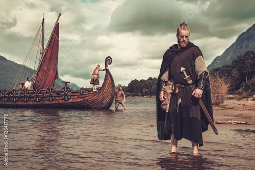 Fotografie, Obraz Viking warrior with sword standing near Drakkar on the seashore