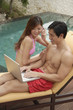 Couple sitting by swimming pool, man using laptop, woman on the phone