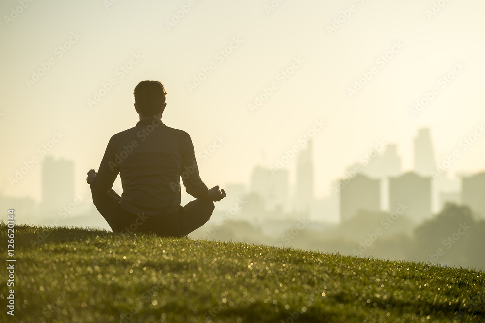 Silhouette of a man sitting in the lotus position meditating on the grassy top of Primrose Hill in front of a misty golden sunrise view of the London city skyline - obrazy, fototapety, plakaty 