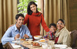 © Alexander - woman stands over dinner table with smiling family